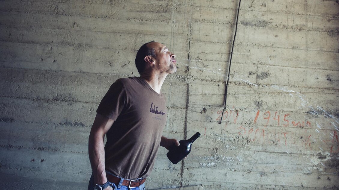 Ein Mann steht in einem Raum mit Betonwänden und schaut nach oben. Er hält eine Flasche in der Hand und zeigt eine nachdenkliche Haltung. | © Weingut Harkamp