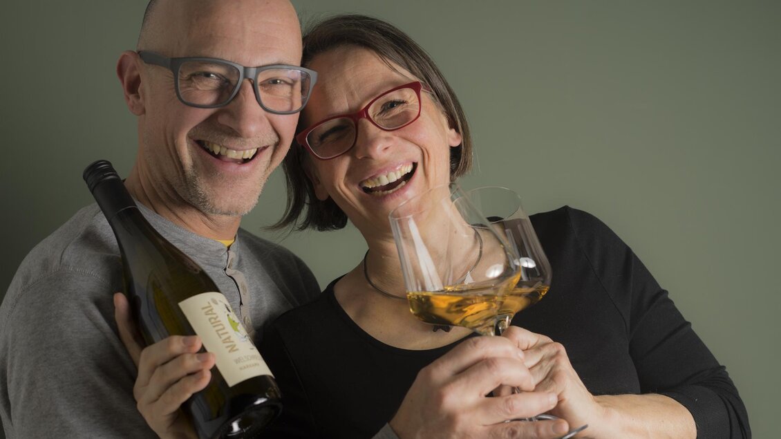 Ein fröhliches Paar lächelt gemeinsam, während es eine Flasche Wein und ein Glas hält. Sie stehen vor einem neutralen, grünen Hintergrund. | © Weingut Harkamp