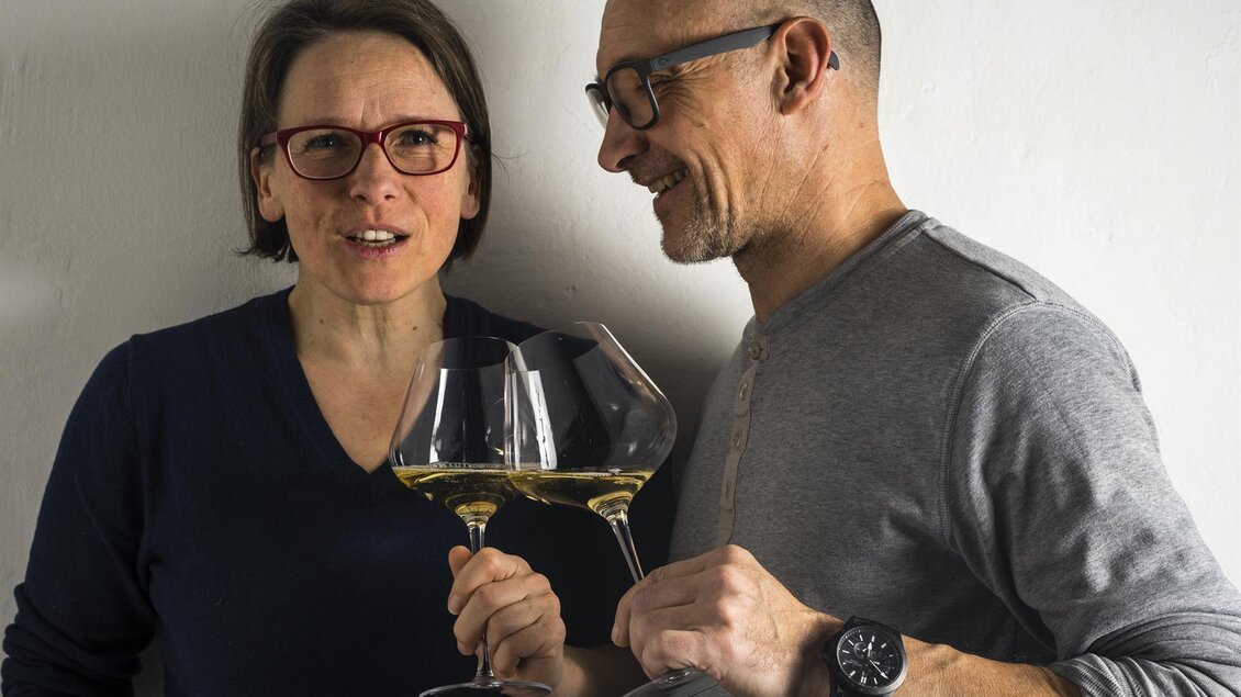 Ein Paar steht lächelnd vor einer neutralen Wand. Beide halten große Gläser mit Wein in der Hand. | © Weingut Harkamp