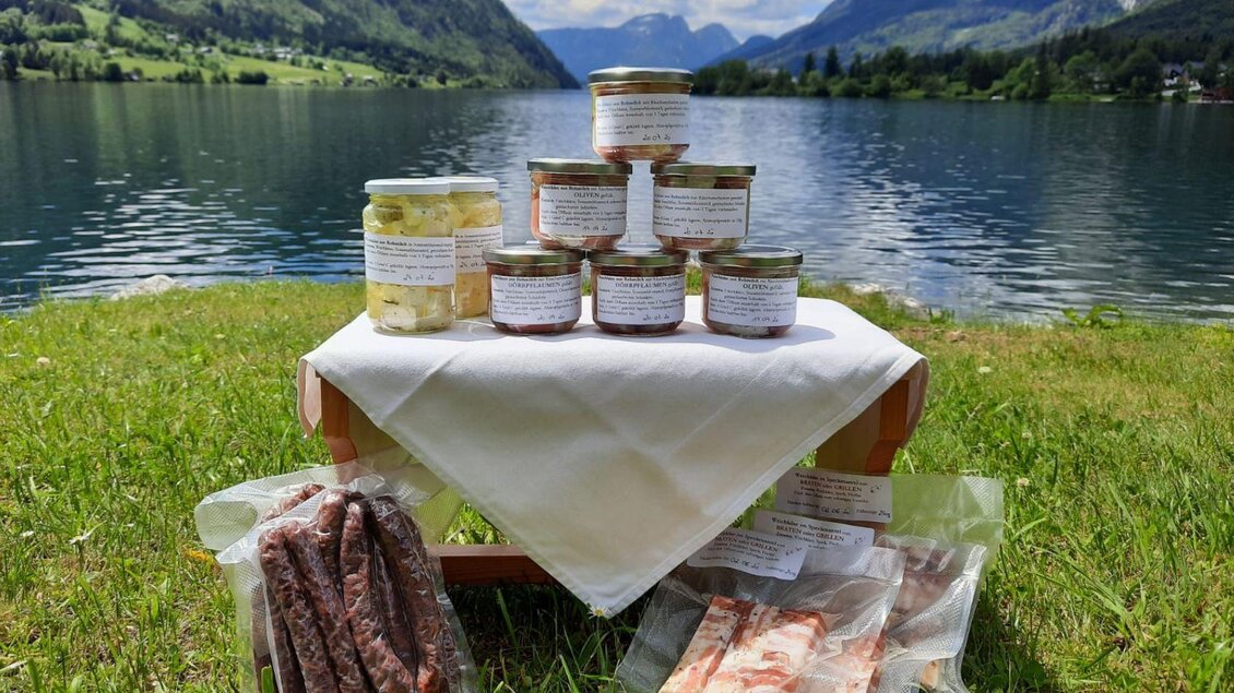 Ein Picknicktisch mit eingelegten Produkten und Wurstwaren steht am Ufer eines klaren Sees. Im Hintergrund sind grüne Berge und ein blauer Himmel zu sehen. | © Bauernhof Butterer
