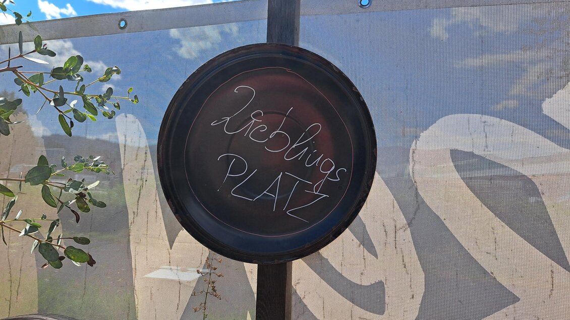 Ein runder Schild mit der Aufschrift "Lieblingsplatz" hängt an einem Holzpfosten. Im Hintergrund ist ein hellblauer Himmel mit weißen Wolken zu sehen. | © TVB_Steininger