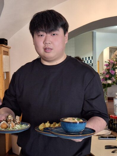 Ein junger Mann steht in einem Restaurant und hält zwei Teller mit Essen. Im Hintergrund sind Tische, Stühle und dekorative Elemente zu sehen. | © leopress