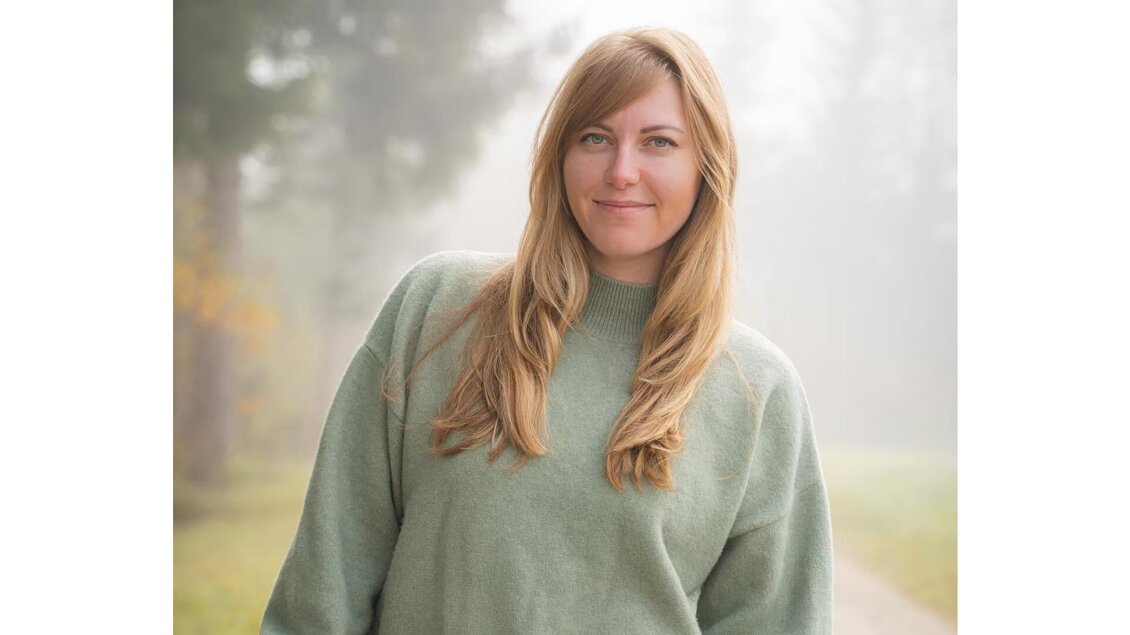 Eine Frau mit langen, blonden Haaren steht in einem nebligen Umfeld. Sie trägt einen grünen Pullover und lächelt sanft in die Kamera. | © Sibel Zechmann Fotografie
