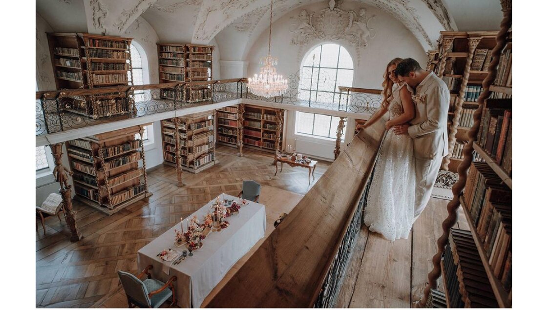 Ein elegantes Paar steht in einer stilvollen Bibliothek mit Bücherregalen. Darunter ist ein festlich gedeckter Tisch zu sehen. | © Sibel Zechmann Fotografie & Content Creation