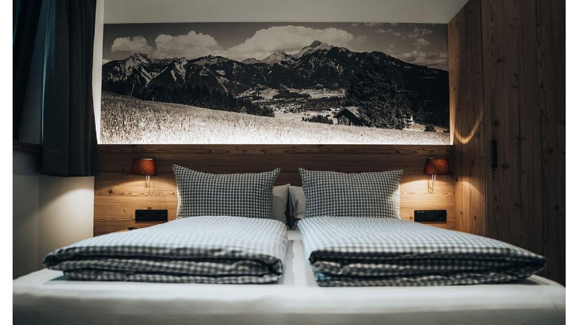 Ein gemütliches Schlafzimmer mit einem großen Bett und karierten Kissen. An der Wand hängt ein beeindruckendes Schwarz-Weiß-Foto von Bergen. | © Sibel Zechmann Fotografie & Content Creation