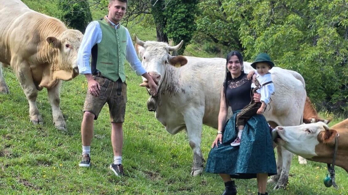 Eine Familie steht auf einer grünen Wiese mit Kühen. Sie sind traditionell gekleidet und lächeln in die Kamera. | © Angelina Skerget