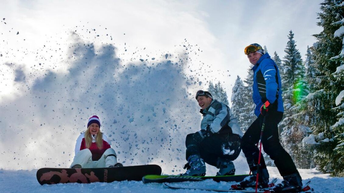 Drei Wintersportler posieren fröhlich im Schnee. Im Hintergrund sind verschneite Bäume und ein strahlend blauer Himmel zu sehen. | © Ikarus | Tom Lamm
