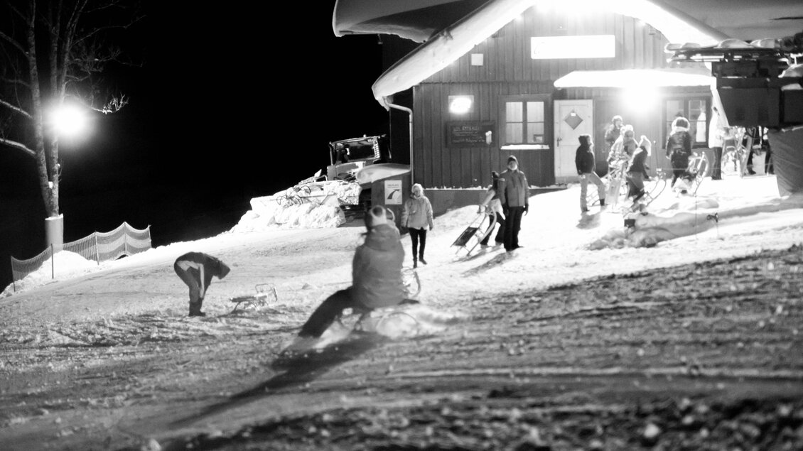 <p>Eine verschneite Landschaft bei Nacht mit Menschen, die zum Rodeln bereit sind. Im Hintergrund sieht man die beleuchtete Hütte am Grafenwiesenlift in Tauplitz, die eine gemütliche Atmosphäre schafft.</p> | © Nico Oussoren, Grafenwiese Tauplitz