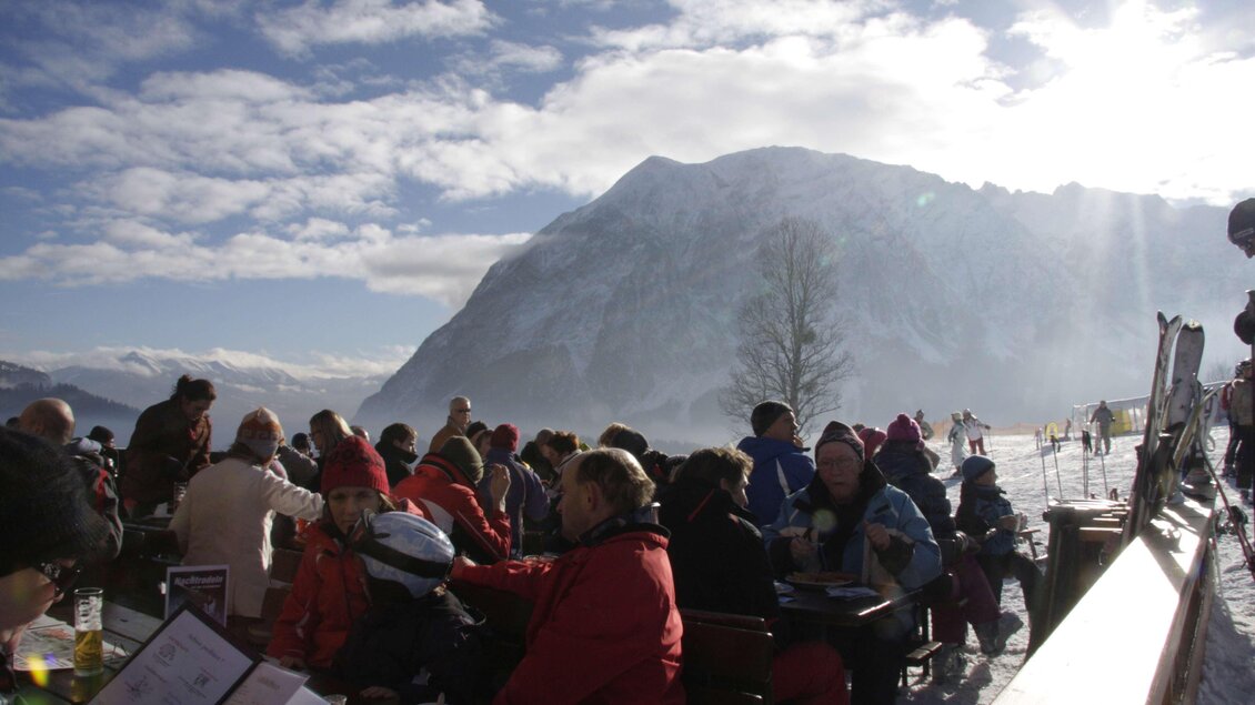 <p>Die belebte Skihütte Grafenwiese mit Menschen, die die Sonne genießen. Im Hintergrund ist der schneebedeckte Grimming zu sehen.</p> | © Grafenwiese Tauplitz