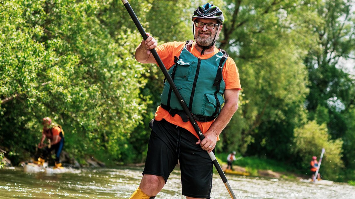 Ein Mann paddelt auf einem SUP-Board in einem ruhigen Gewässer. Im Hintergrund sind Bäume und weitere Personen zu sehen, die aktiv sind. | © Robert und Verena Koch