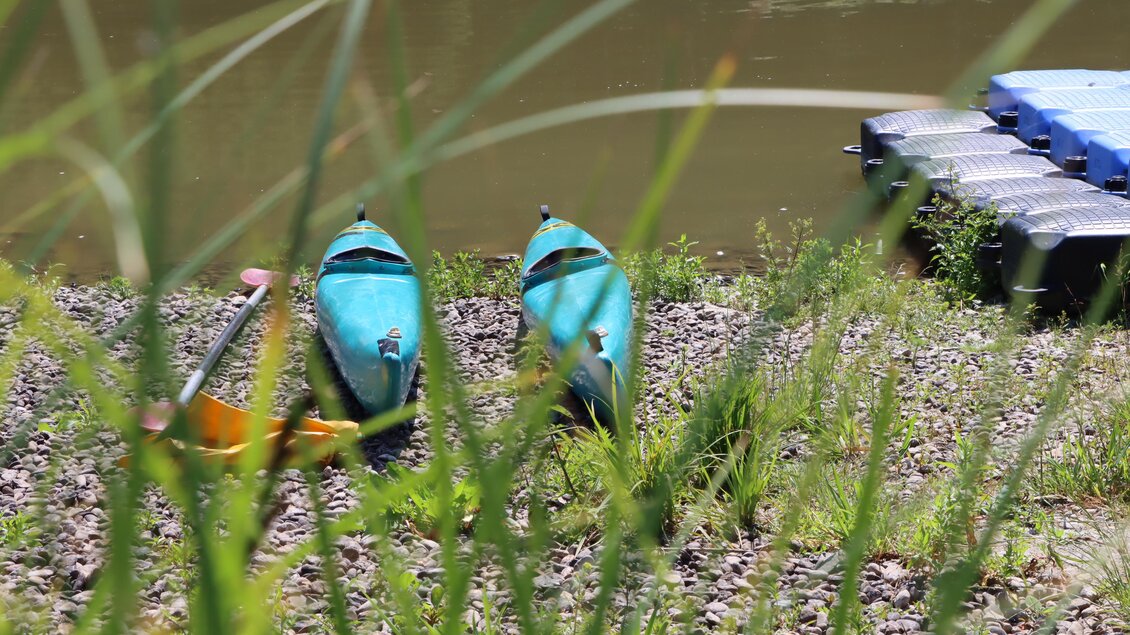 Zwei Kajaks stehen am Ufer eines Gewässers, umgeben von Gras und Steinen. Im Hintergrund sieht man einen schwimmenden Steg. | © TVB Erzberg-Leoben