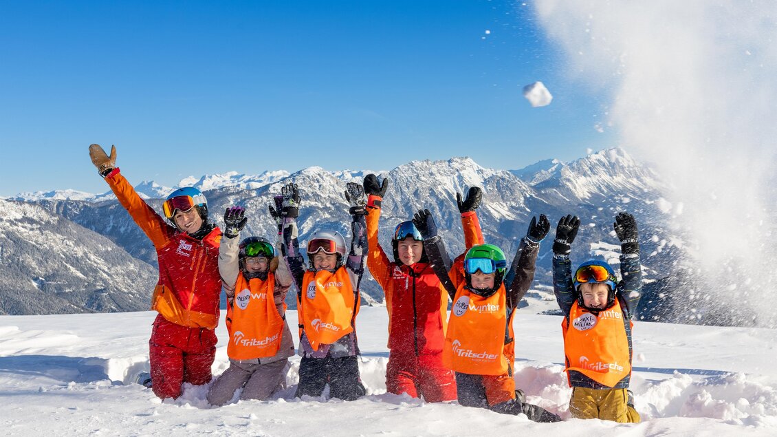 Skilehrerin und Kinder in orangefarbenen Westen knien im Tiefschnee und strecken jubelnd die Arme nach oben; im Hintergrund ein verschneites Bergpanorama unter blauem Himmel. | © Skischule Tritscher