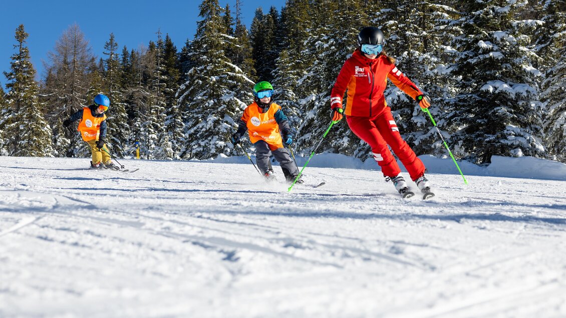 Skilehrerin fährt mit zwei Kindern schwungvoll über eine Piste; alle tragen Helme und orangefarbene Westen, verschneiter Wald im Hintergrund. | © Skischule Tritscher