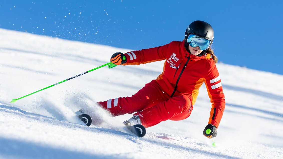 Skilehrerin in roter Skischulkleidung fährt eine saubere Carving-Kurve, grüner Skistock und aufspritzender Schnee setzen Dynamik vor blauem Himmel. | © Skischule Tritscher