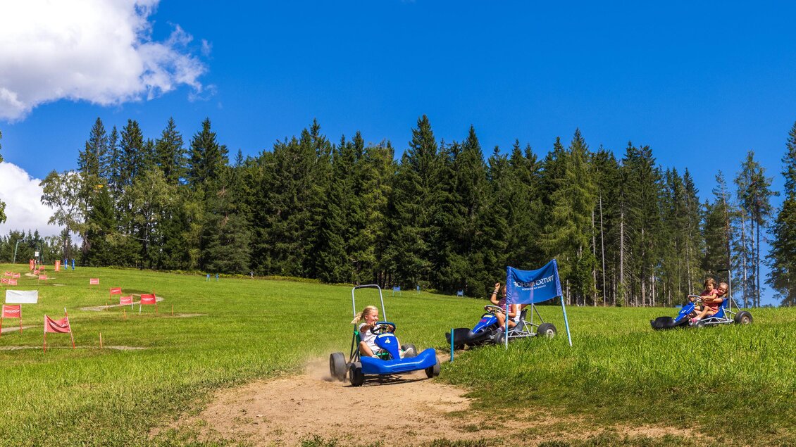 Zwei Personen fahren in blauen Rutschautos auf einer grünen Wiese. Im Hintergrund sind Bäume und ein klarer blauer Himmel zu sehen. | © Hans-Peter Steiner