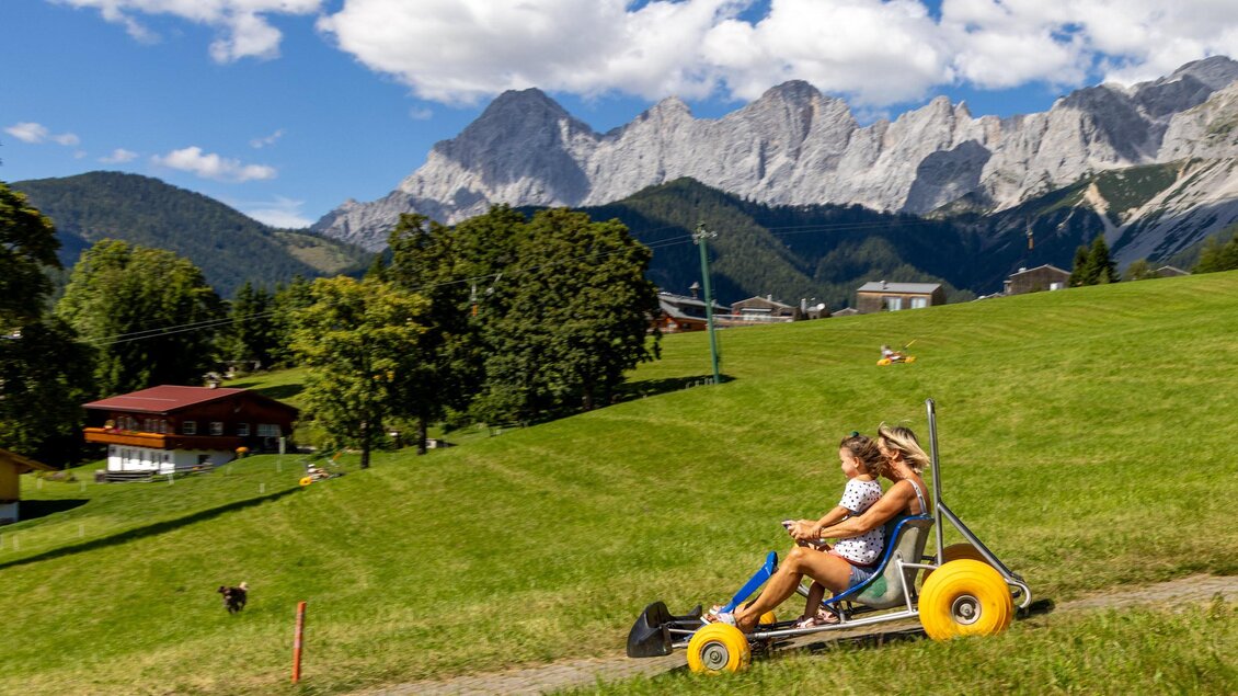 Zwei Personen fahren mit einem Go-Kart einen grünen Hang hinunter. Im Hintergrund sind Berge und Bäume zu sehen. | © Hans-Peter Steiner