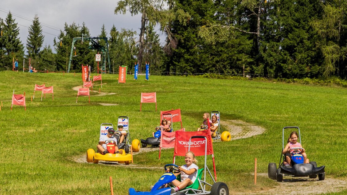 Kinder fahren mit Karts auf einer kleinen Strecke in einer grünen Landschaft. Im Hintergrund sind Bäume und ein blauer Himmel zu sehen. | © Hans-Peter Steiner