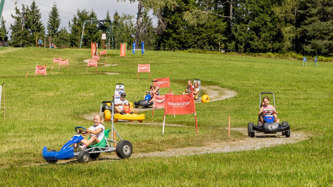 Kinder fahren mit Karts auf einer Strecke in einem grünen Gelände. Im Hintergrund sind Bäume und Sicherheitsmarkierungen zu sehen. | © Hans-Peter Steiner