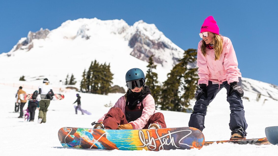Zwei Snowboarderinnen im Schnee, eine sitzt auf ihrem Board und die andere steht daneben in winterlicher Kleidung. Im Hintergrund sind verschneite Berge und weitere Snowboarder zu sehen. | © Lib Tech