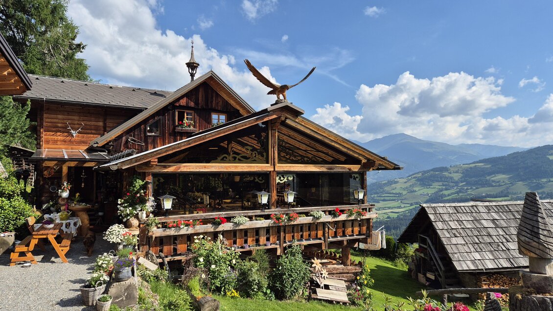 Ein gemütliches Holzhaus mit einem großen Balkon und bunten Blumen. Im Hintergrund erstrecken sich malerische Berge unter einem blauen Himmel. | © TV Murau