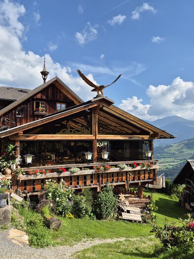 Ein gemütliches Holzhaus mit einem großen Balkon und bunten Blumen. Im Hintergrund erstrecken sich malerische Berge unter einem blauen Himmel. | © TV Murau