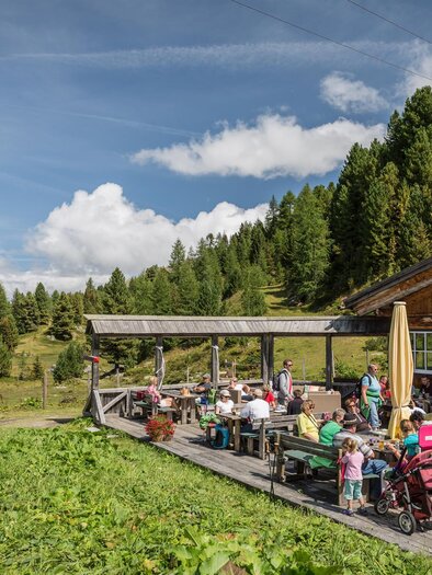 Eine Almhütte mit Außenterrasse, auf der viele Gäste sitzen und essen. Im Hintergrund sind grüne Wiesen und Wälder zu sehen. | © TMG Turracher Höhe Marketing GmbH