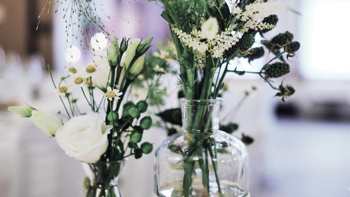 Zwei elegante Blumenvasen auf einem Holzbrett. Sie enthalten frische, weiße Blumen und grünes Blattwerk. | © Sorgerhof