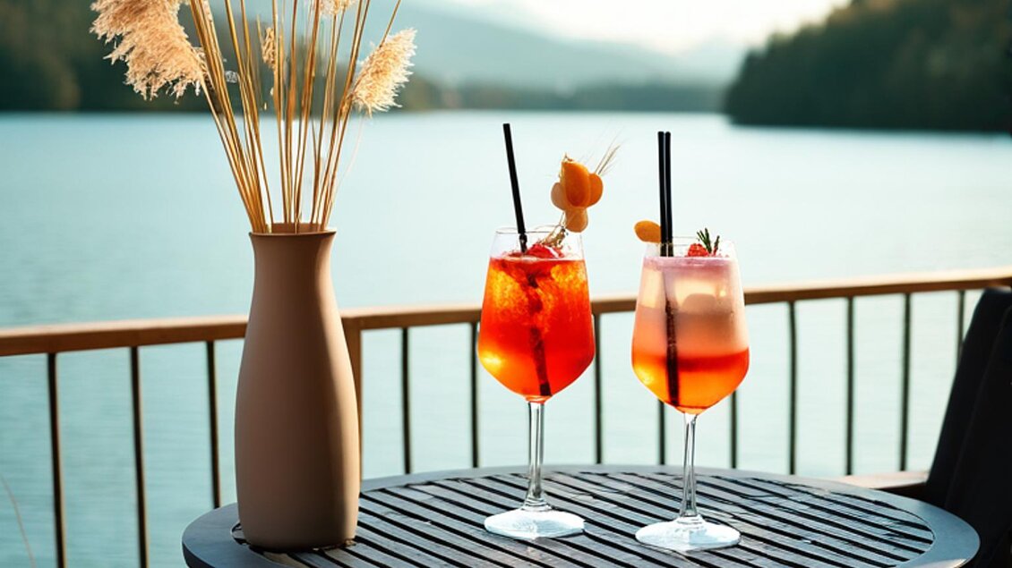 Ein schöner Tisch auf einer Terrasse am Wasser mit zwei bunten Cocktails. Im Hintergrund sieht man Berge und einen klaren Himmel. | © Holzerbauer