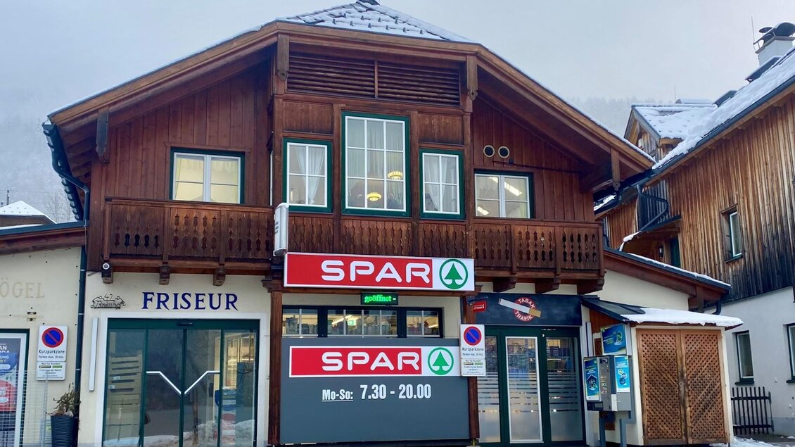 Ein traditionelles Holzgebäude mit einem Spar-Markt und einem Friseursalon. Die Fassade hat große Fenster und ist in einer ländlichen Umgebung gelegen. | © Bettina Scheck