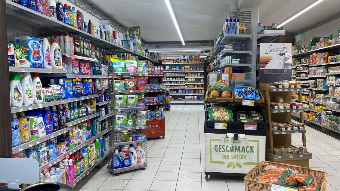 Ein gut sortierter Supermarkt mit Regalen voller Reinigungsprodukte und Lebensmittel. Auf einem Tisch sind frische Produkte und eine Werbetafel zu sehen. | © Bettina Scheck