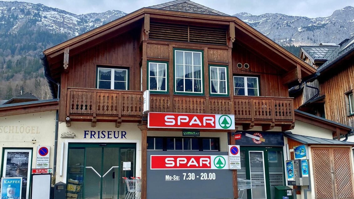 Ein traditionelles Holzgebäude mit einem SPAR-Laden im Erdgeschoss und einem Friseur. Im Hintergrund sind schneebedeckte Berge sichtbar. | © Bettina Scheck