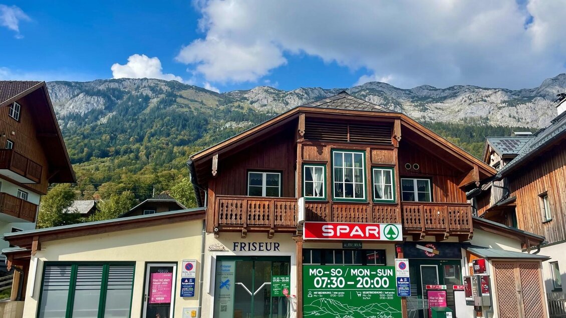 Ein traditionelles Gebäude mit Holzverkleidung, das einen Friseur und einen Supermarkt zeigt. Im Hintergrund sind majestätische Berge und ein blauer Himmel zu sehen. | © Bettina Scheck