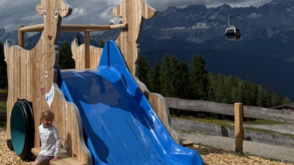 Ein Spielplatz mit einer blauen Rutsche, die von zwei Ziegenfiguren umgeben ist. Im Hintergrund sind Berge und eine Seilbahn zu sehen. | © Reiteralm