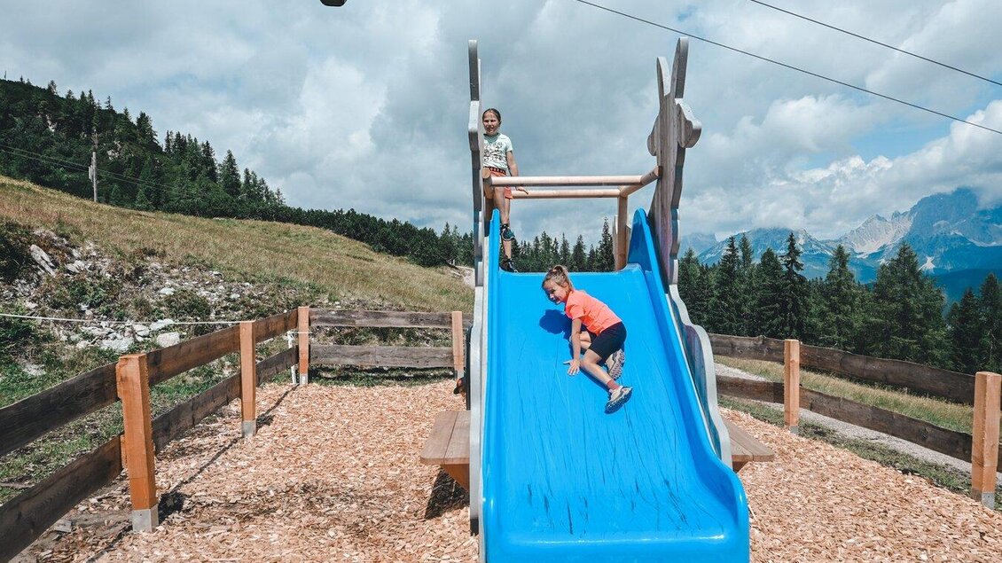 Ein Spielplatz in den Bergen mit einer blauen Rutsche. Zwei Kinder spielen draußen bei schönem Wetter. | © Reiteralm