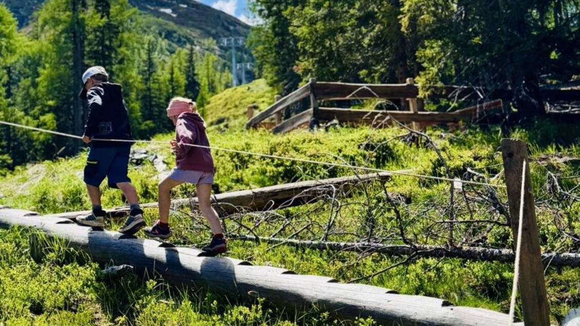 Zwei Kinder überqueren eine Holzbrücke in einer malerischen Landschaft. Im Hintergrund sind Bäume und Berge sichtbar, während die Sonne scheint. | © Reiteralm