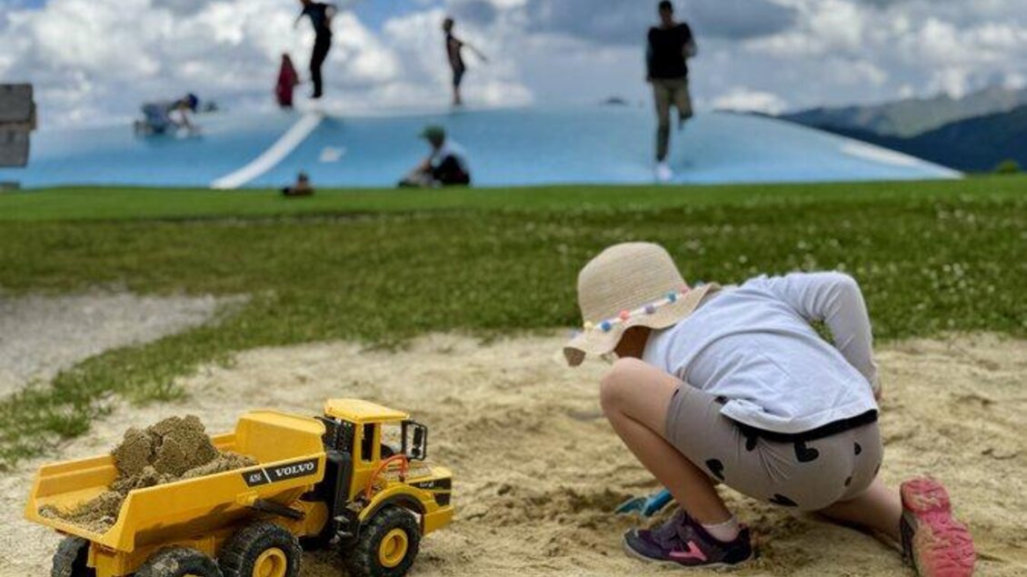 Ein Kind spielt im Sand mit einem Spielzeug-Lkw. Im Hintergrund sind Menschen, die auf einem Hügel spielen. | © Reiteralm