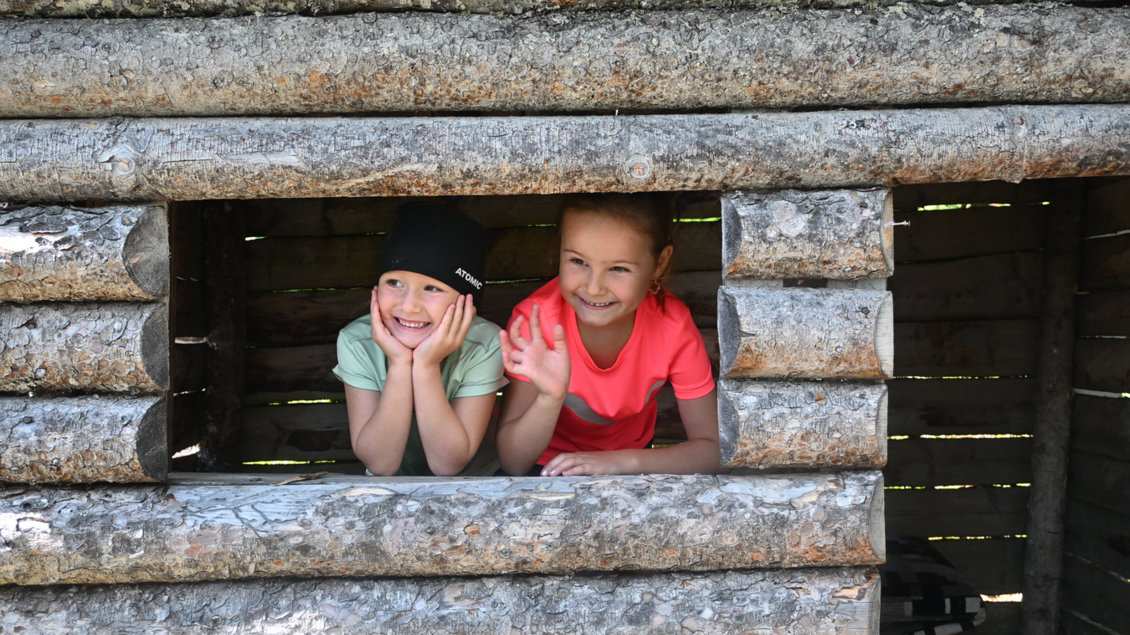 Zwei Mädchen schauen neugierig aus einem Holzhaus. Sie lächeln und wirken glücklich. | © Reiteralm