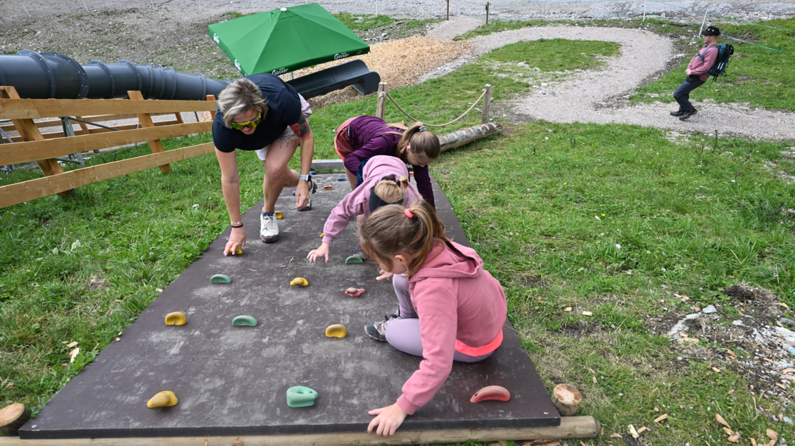 Kinder klettern begeistert an einer Kletterwand auf einem Spielplatz. Im Hintergrund ist ein Erwachsener zu sehen, der ihnen hilft. | © Reiteralm