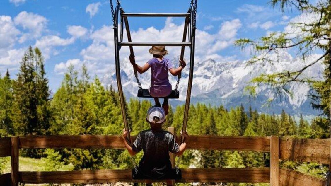 Zwei Kinder schwingen auf einer Schaukel mit Blick auf die Berge. Der Himmel ist blau und es gibt viele Bäume in der Umgebung. | © Reiteralm