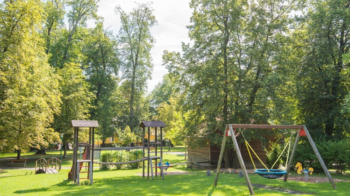 Ein Spielplatz in einem Park mit grünen Wiesen und hohen Bäumen. Es gibt Schaukeln und Klettergerüste zum Spielen. | © Helmut Schweighofer