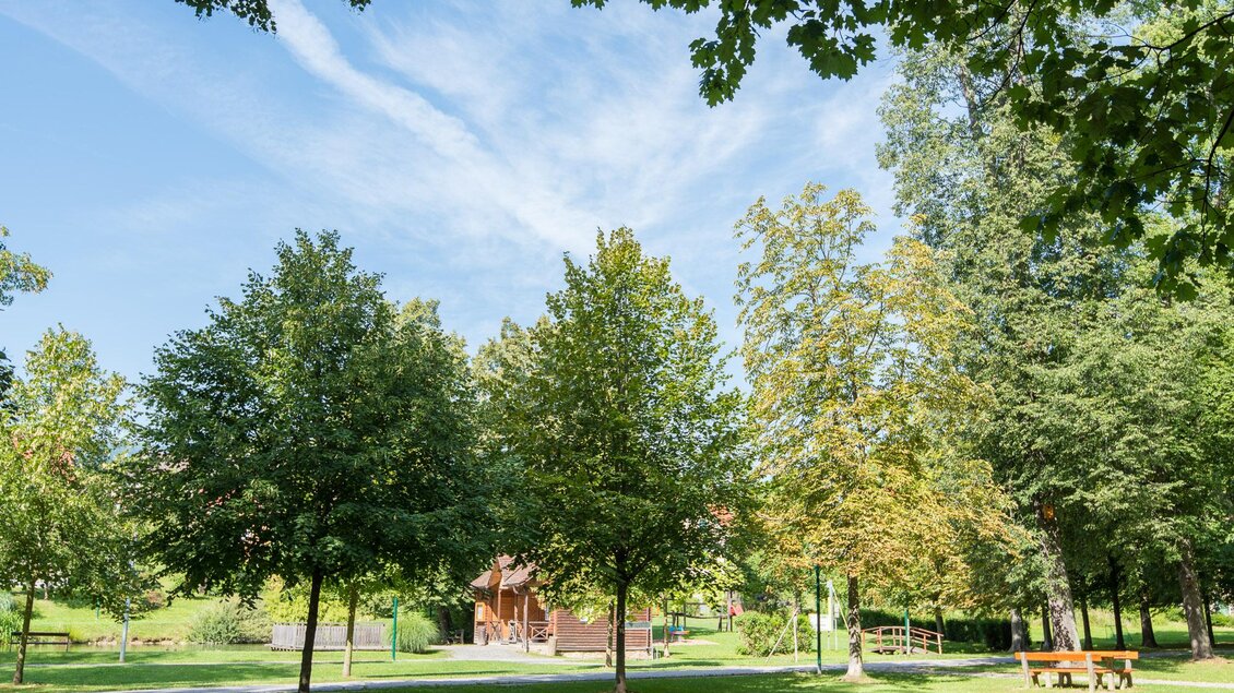 Ein ruhiger Park mit grünen Bäumen und einem blauen Himmel. Im Hintergrund ist ein Gebäude zu sehen. | © Helmut Schweighofer