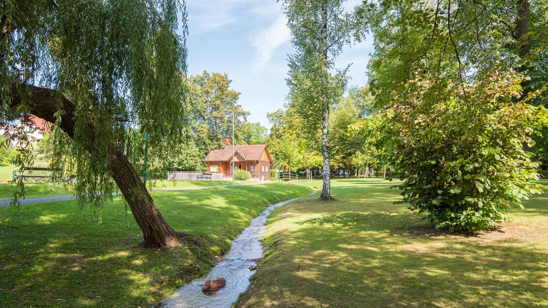 Ein malerischer Garten mit einem kleinen Bach, umgeben von Bäumen und Wiesen. Im Hintergrund ist ein rotes Haus sichtbar. | © Helmut Schweighofer