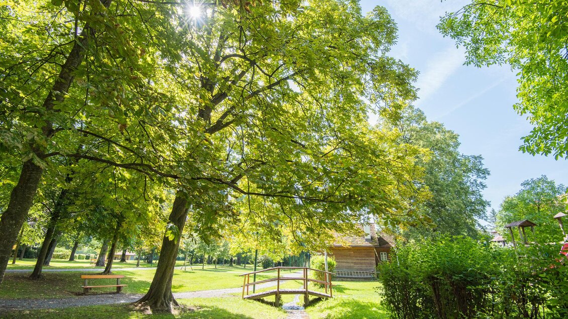 Ein schöner Park mit vielen grünen Bäumen und viel Sonnenlicht. Im Hintergrund ist eine kleine Brücke und ein Holzhaus zu sehen. | © Helmut Schweighofer
