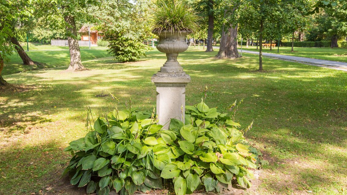 Ein schöner Garten mit üppigen Pflanzen und einem dekorativen Blumentopf. Im Hintergrund sind Bäume und ein Pfad zu sehen. | © Helmut Schweighofer