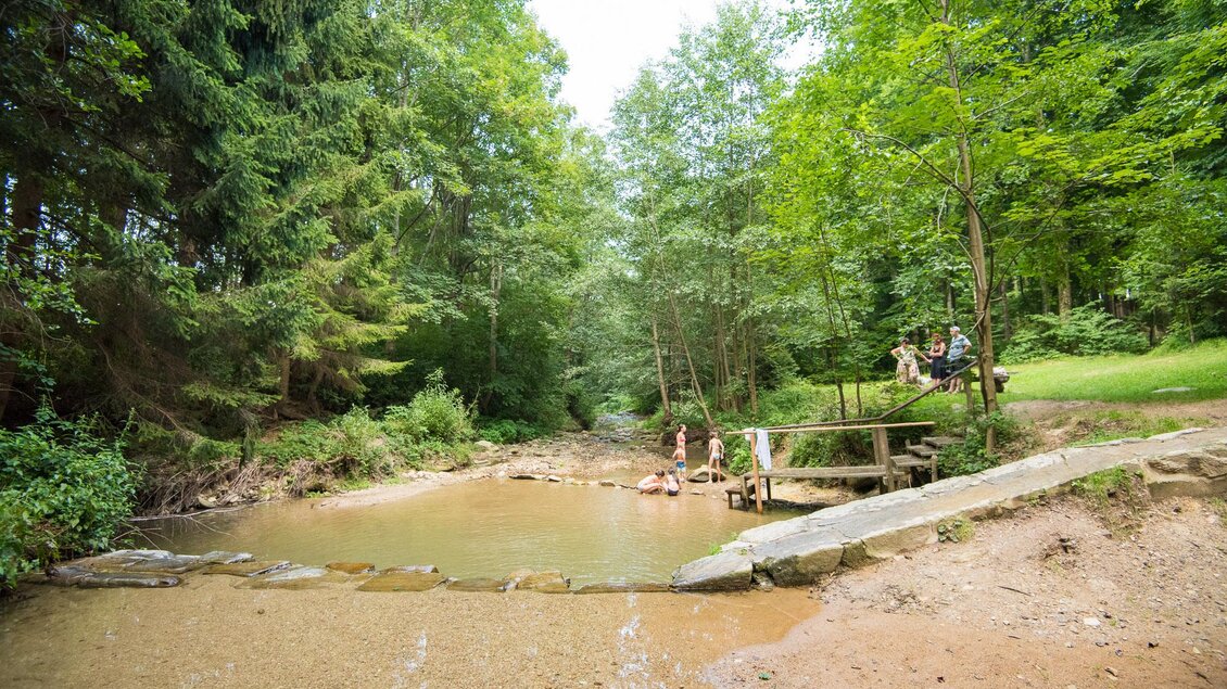 Ein ruhiger Bach umgeben von grünem Wald. Menschen entspannen am Ufer und genießen die Natur. | © Helmut Schweighofer