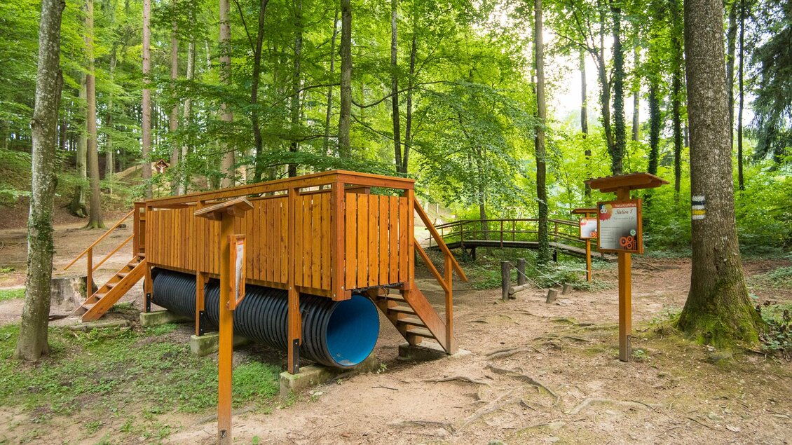 Ein Spielplatz aus Holz im Wald, umgeben von grünen Bäumen. Die Struktur besteht aus einer Spielplattform mit einer Rutsche und anderen Spielgeräten. | © Helmut Schweighofer
