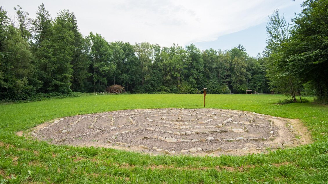 Ein spiralförmiges Steine-Labyrinth in einer grünen Wiese, umgeben von Bäumen. Der Himmel ist bewölkt und die Atmosphäre wirkt ruhig und naturverbunden. | © Helmut Schweighofer
