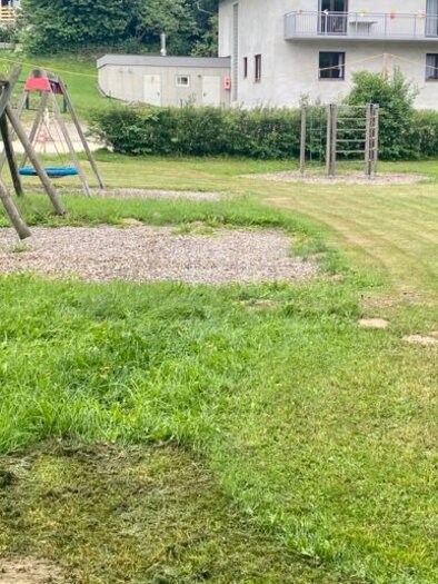 Ein Spielplatz mit Schaukeln und einer Rasenfläche. Im Hintergrund sieht man ein Gebäude und einige Spielmöglichkeiten. | © Emily Cividino
