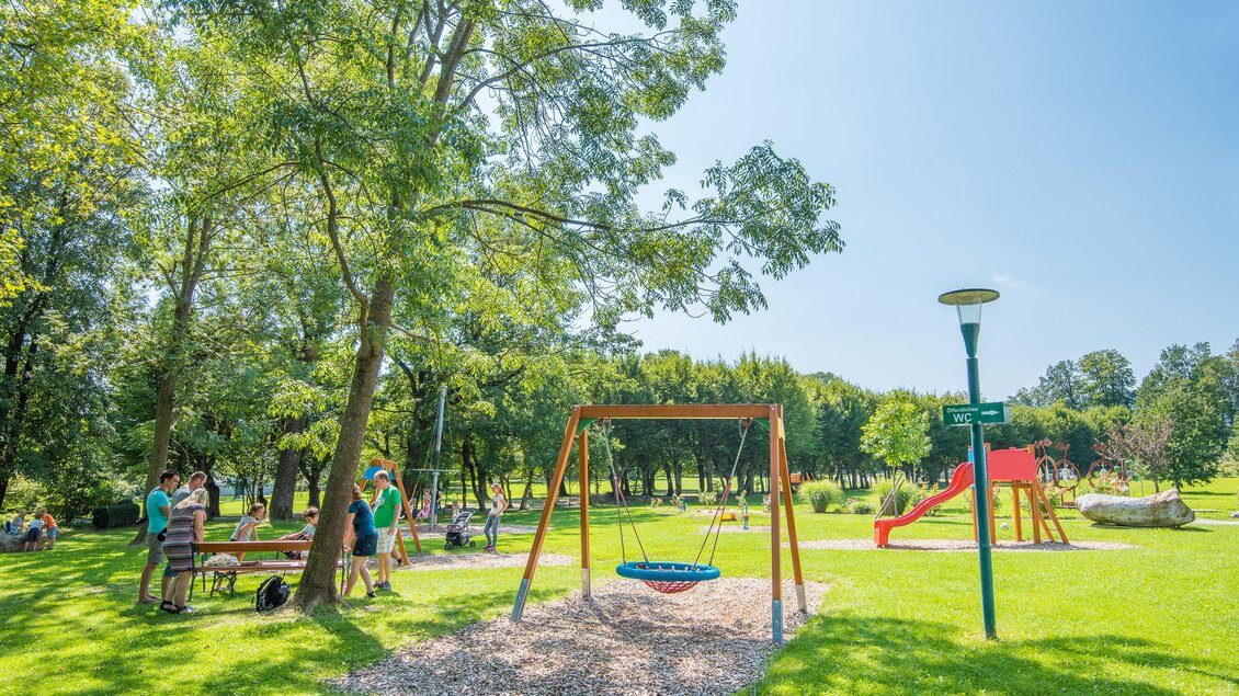 Ein schöner Spielplatz im Park mit einer Schaukel und einer Rutsche. Umgeben von Bäumen und viel grünem Gras, genießen Familien das sonnige Wetter. | © Helmut Schweighofer