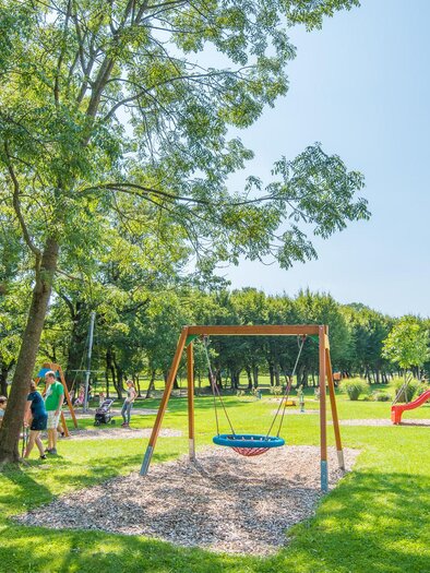Ein schöner Spielplatz im Park mit einer Schaukel und einer Rutsche. Umgeben von Bäumen und viel grünem Gras, genießen Familien das sonnige Wetter. | © Helmut Schweighofer